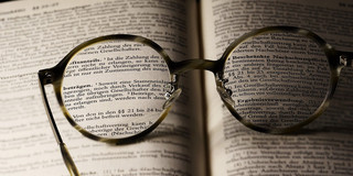 Eine Nahaufnahme von Brillengläsern, die auf einer geöffneten Buchseite liegen. Der Text in den Gläsern wird klarer sichtbar, während der Hintergrund verschwommen bleibt. Die Brille symbolisiert das Streben nach Wissen und Verständnis, während die gedruckten Wörter im Buch auf juristische oder geschäftliche Themen hinweisen. Diese Komposition vermittelt den Eindruck von Neugier und dem Wunsch, tiefere Einsichten zu gewinnen.