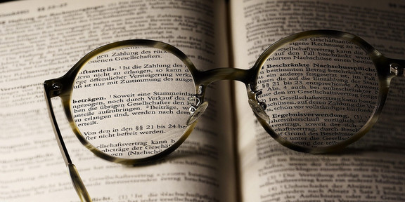 Eine Nahaufnahme von Brillengläsern, die auf einer geöffneten Buchseite liegen. Der Text in den Gläsern wird klarer sichtbar, während der Hintergrund verschwommen bleibt. Die Brille symbolisiert das Streben nach Wissen und Verständnis, während die gedruckten Wörter im Buch auf juristische oder geschäftliche Themen hinweisen. Diese Komposition vermittelt den Eindruck von Neugier und dem Wunsch, tiefere Einsichten zu gewinnen.