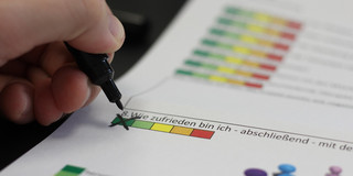 Eine Hand hält einen schwarzen Marker und zeichnet ein X auf ein Dokument, das eine Bewertungsskala zeigt. Die Skala ist in den Farben Grün, Gelb und Rot unterteilt und steht unter der Frage: "8. Wie zufrieden bin ich - abschließend - mit der..." Im Hintergrund sind grafische Darstellungen von Personen in einem Meeting zu sehen.