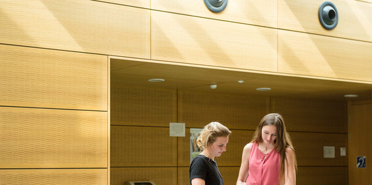 Studentinnen mit Buch am Geländer Zwei Studentinnen stehen mit einem Buch am Geländer.
