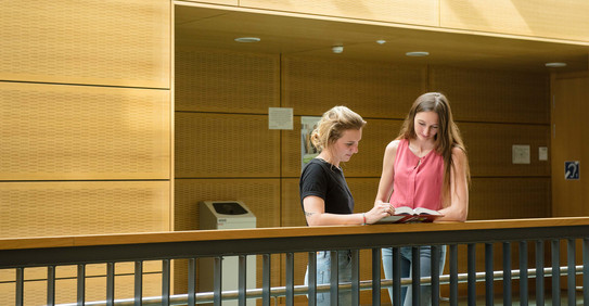 Studentinnen mit Buch am Geländer Zwei Studentinnen stehen mit einem Buch am Geländer.